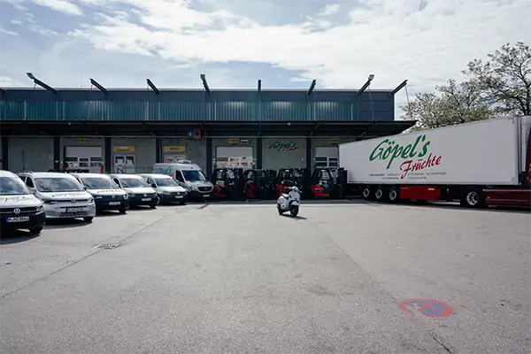 Moderne Lagerhalle mit Laderampe. Davor 6 kleine PKW-Transporter, 4 Gabelstapler und ein Sattelzug LKW mit Göpels-Aufschrift. In der Mitte eine weiße Vespa.