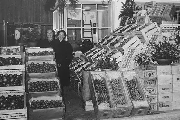 Schwarz-Weiß-Aufnahme eines Obst-Standes in den 1950er Jahren. Zwischen verschiedenen Obstkisten stehen eine junge Frau und ein Mann